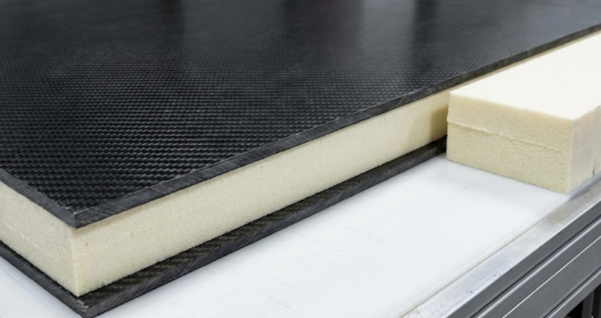 A large carbon fiber composite panel with a foam core displayed next to a raw block of foam material.