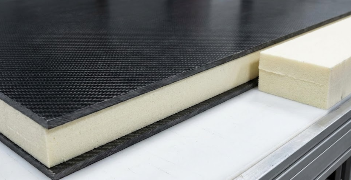 A large carbon fiber composite panel with a foam core displayed next to a raw block of foam material.
