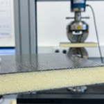 A close-up side view of a carbon fiber foam sandwich sample held in front of a mechanical testing machine.