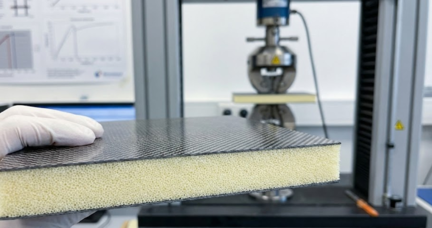 A close-up side view of a carbon fiber foam sandwich sample held in front of a mechanical testing machine.