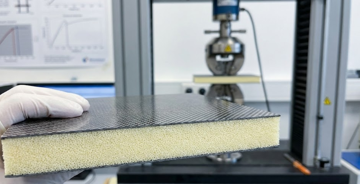 A close-up side view of a carbon fiber foam sandwich sample held in front of a mechanical testing machine.
