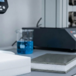 A laboratory setting featuring a microscope, chemical glassware, and samples of white foam and finished composite panels.